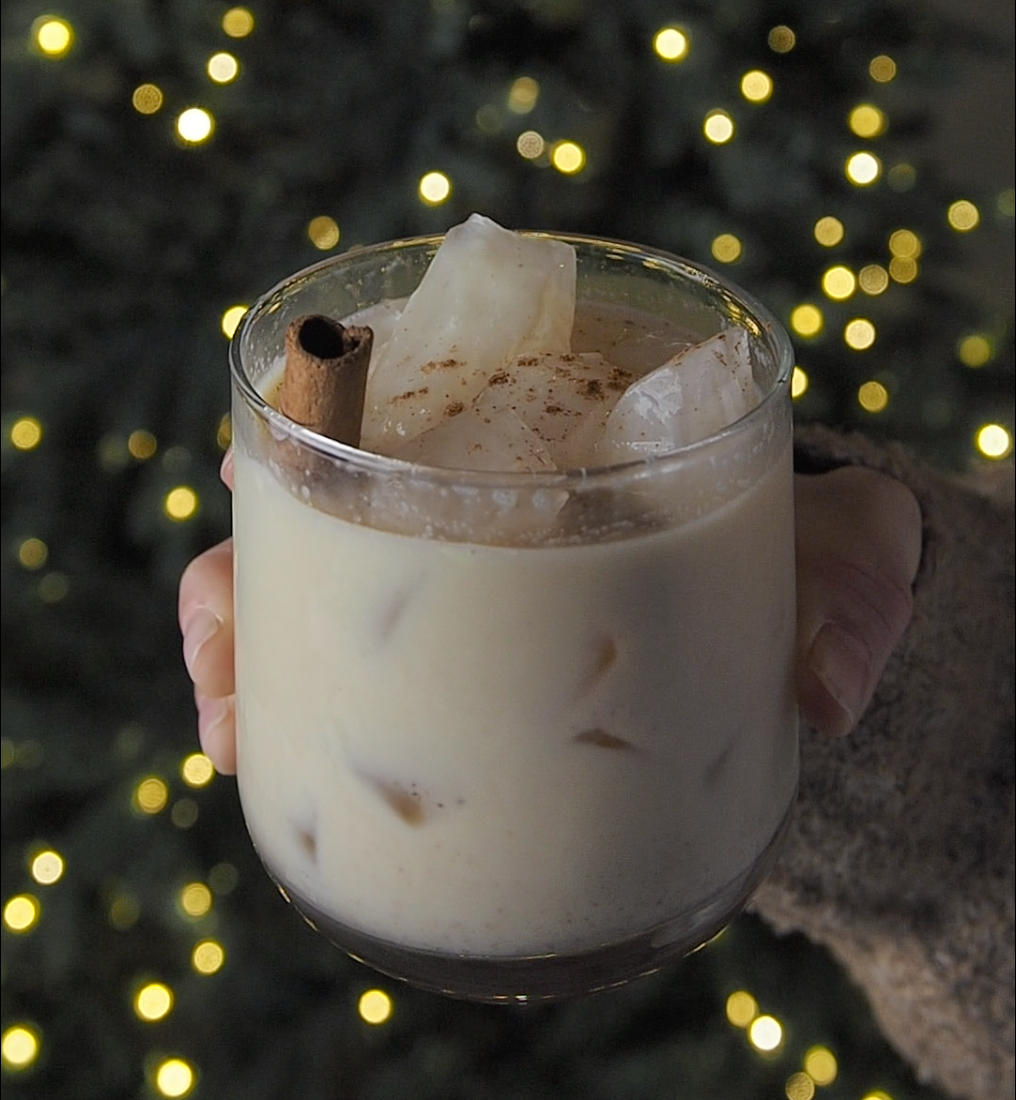 Glass of Bee Mafia Honey Eggnog glass with ice & cinnamon stick held by hand in front of christmas tree.